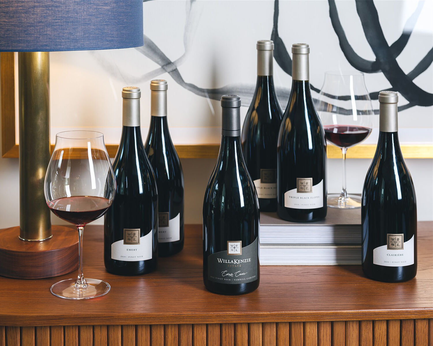 Six bottles of wine laying on wooden furniture next to books, a lamp and two glasses of red wine.